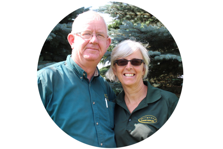 Two people wearing dark green shirts stand side-by-side and smile in front of evergreen trees inside a circular frame.