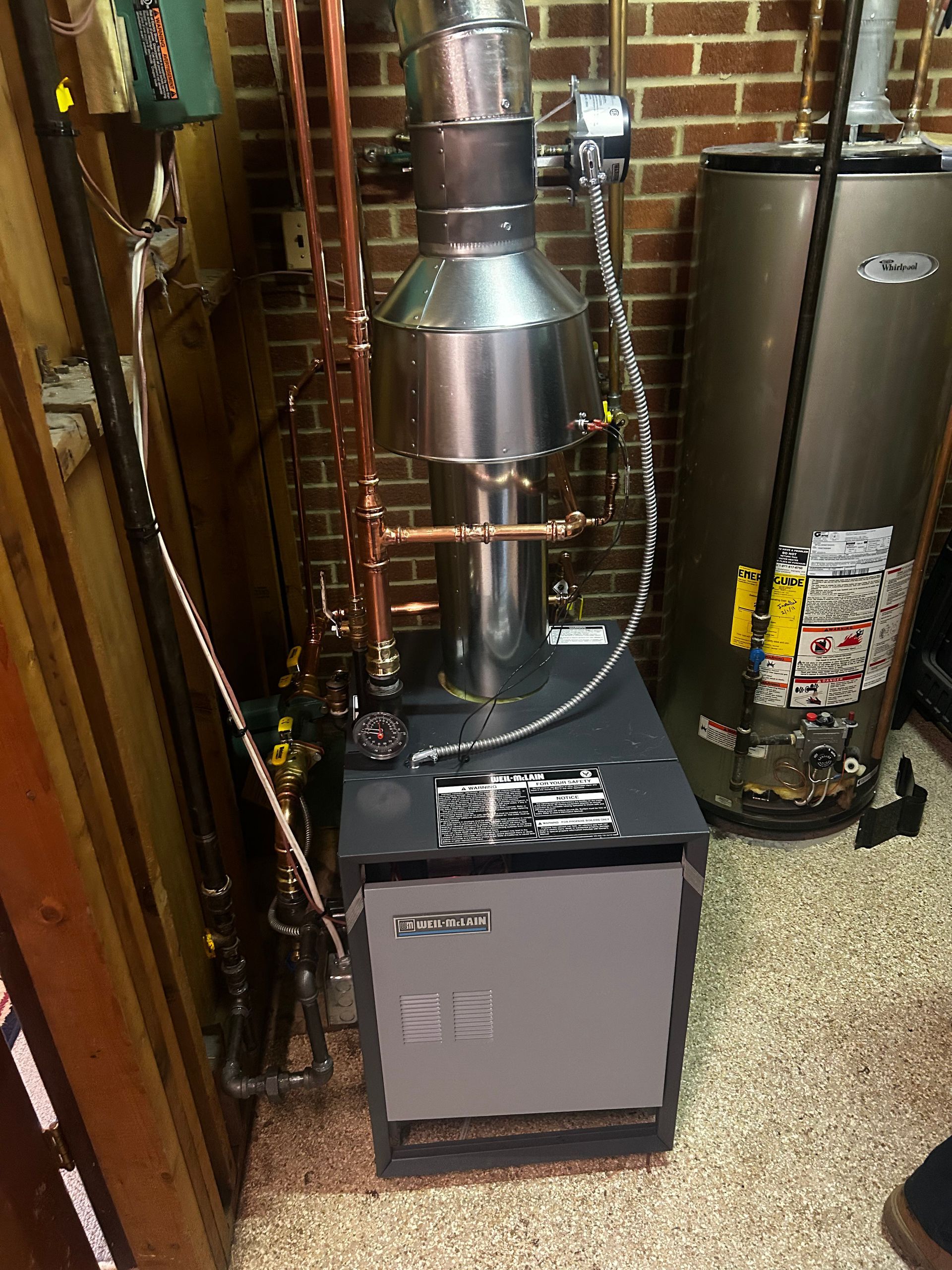 A residential heating system with a metal exhaust stack and copper piping is installed against a brick wall, next to a water heater.