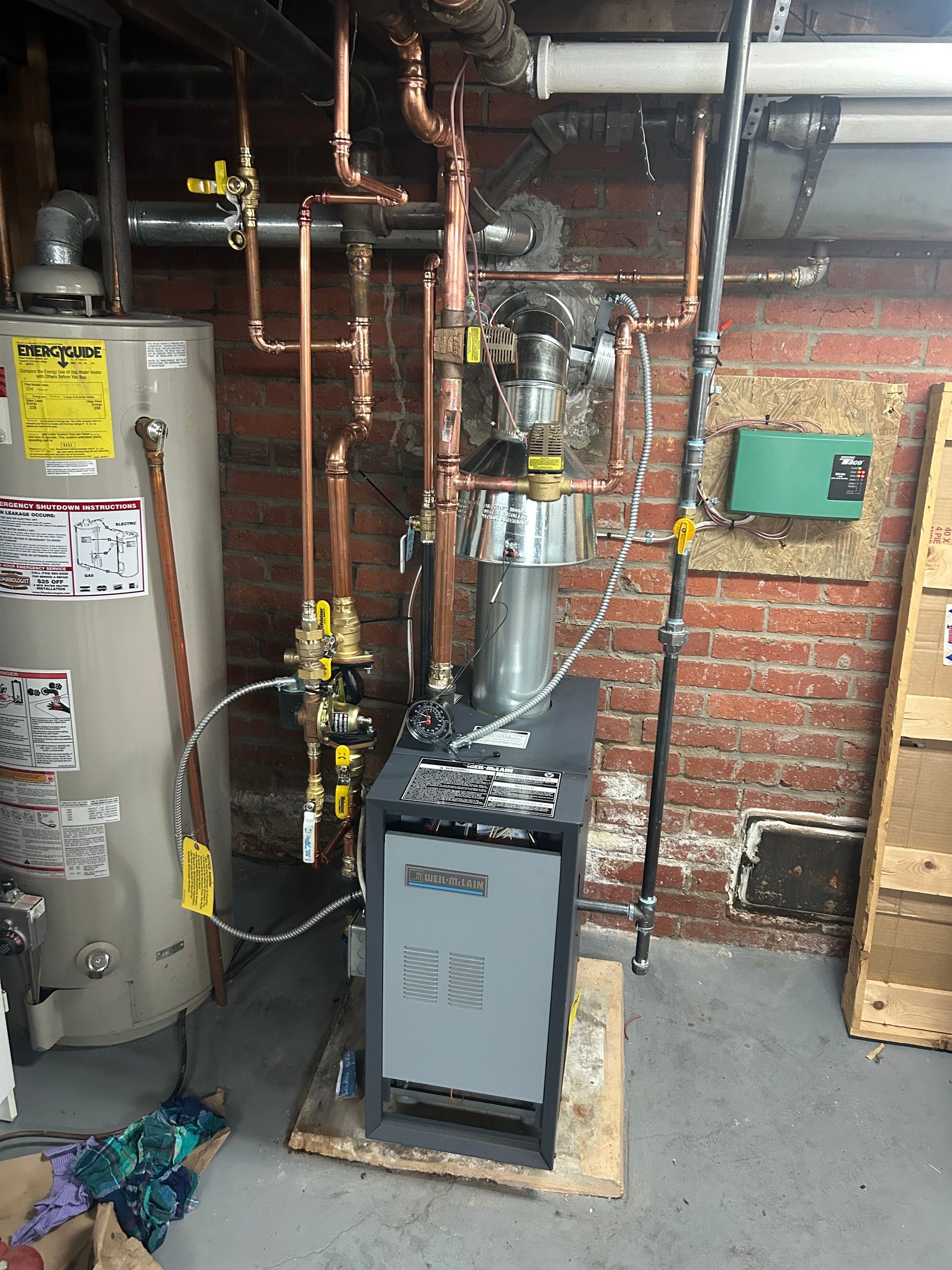 A gray heating system and a water heater with copper pipes in a utility room with exposed brick.