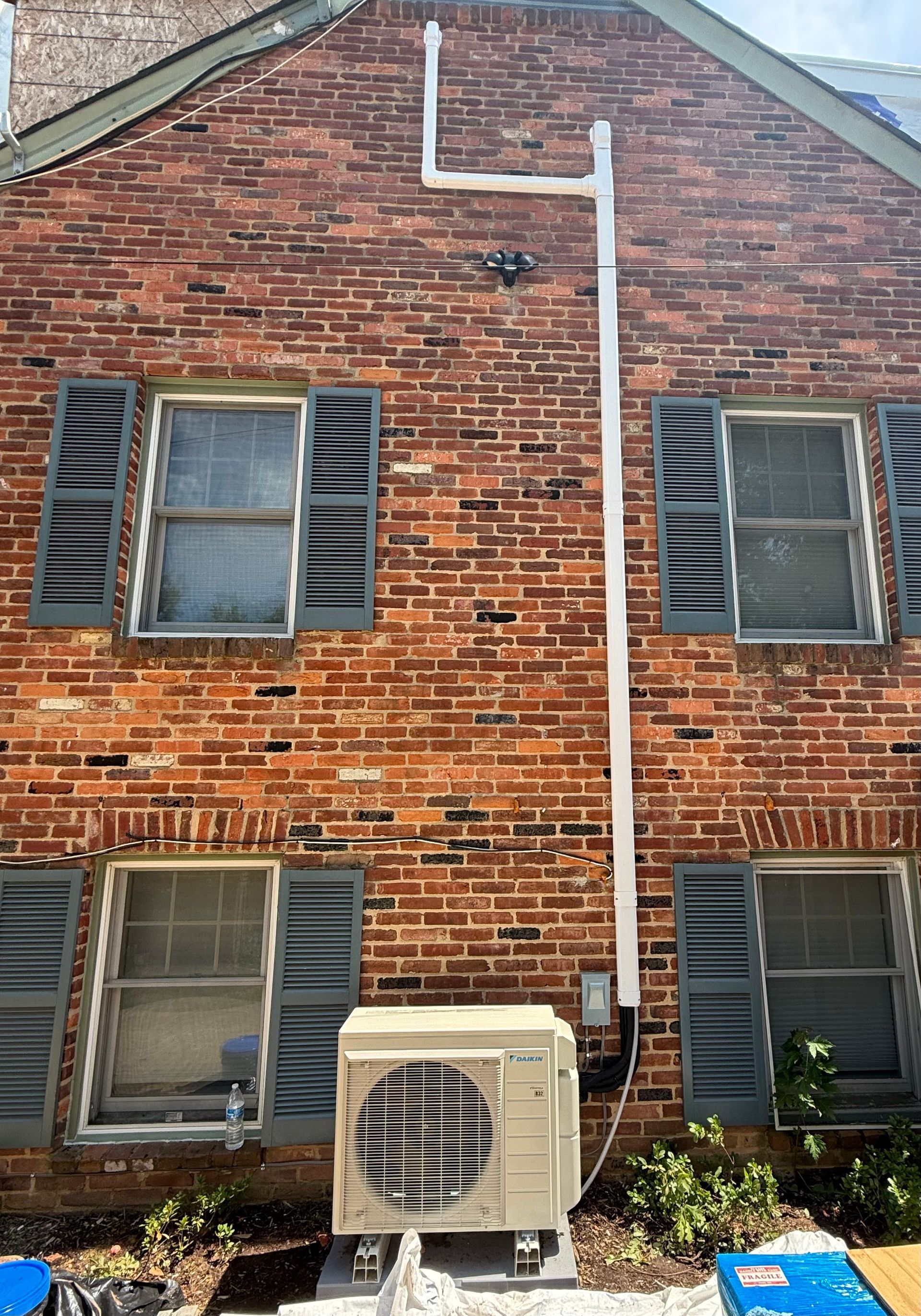 A brick building exterior with gray shutters and an air conditioning unit below the windows. White piping runs up the wall.