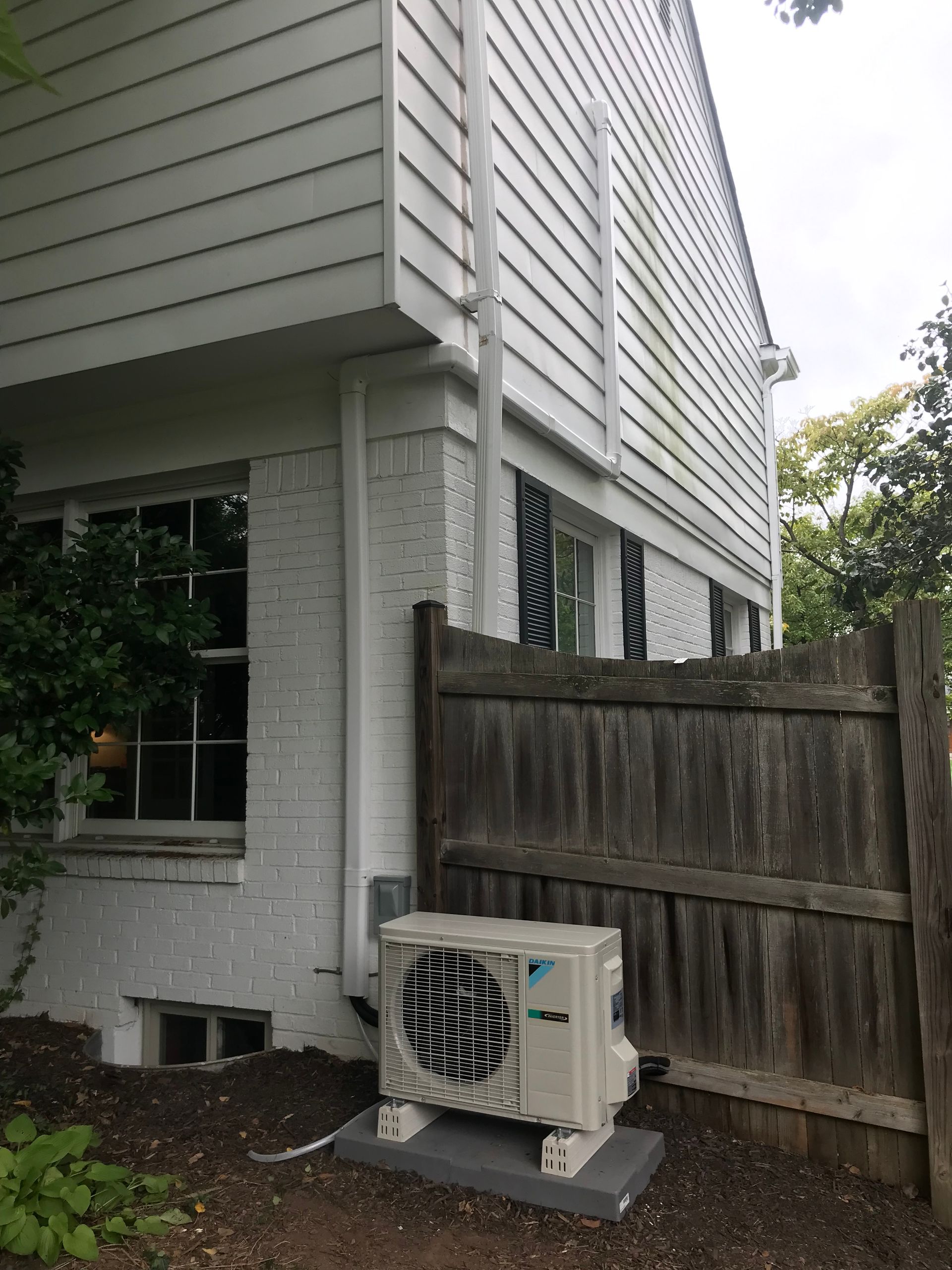 Air conditioning unit mounted on a concrete slab next to a white brick house with a wooden fence in a yard.