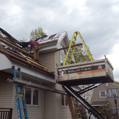 Roofers working on a house with a lift; gray siding, green lawn, clear sky.