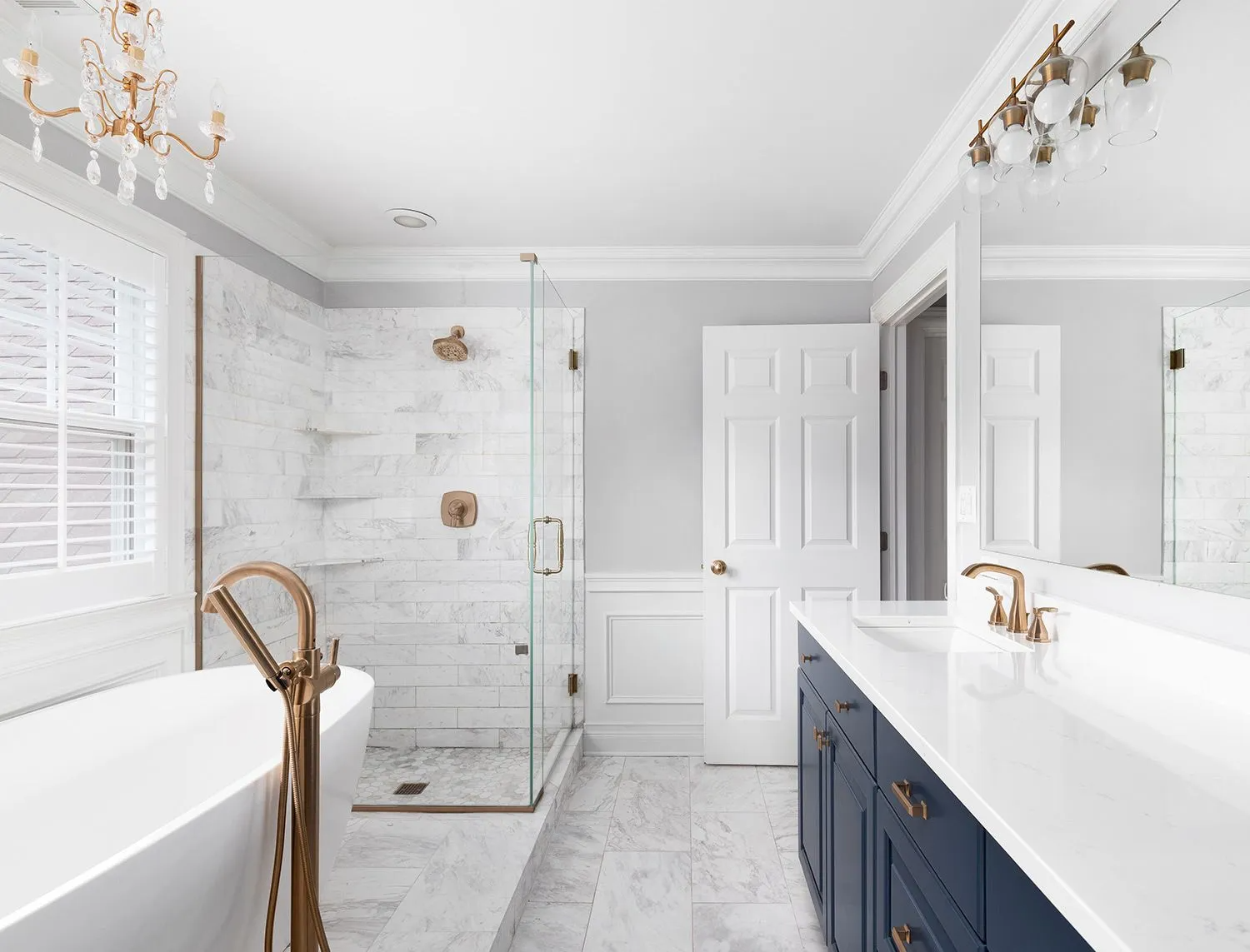 Elegant white bathroom with marble shower, blue vanity, gold fixtures, and chandelier.
