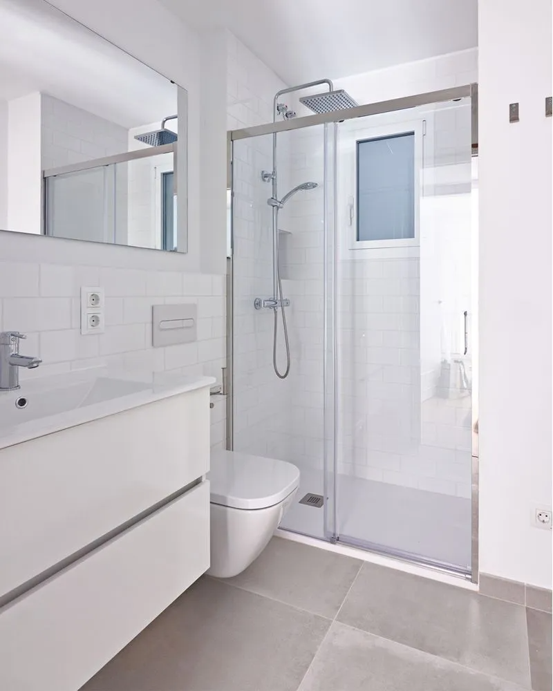 White bathroom with a shower, toilet, and sink. Grey floor tiles, a window, and a mirror are present.