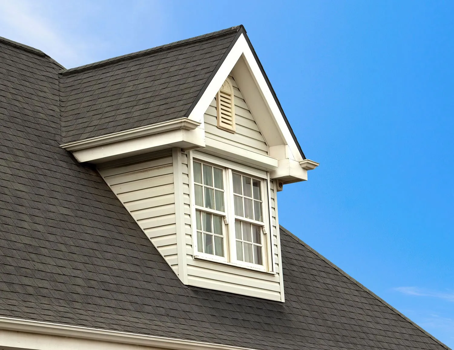 Dormer window on a dark gray shingled roof against a bright blue sky.