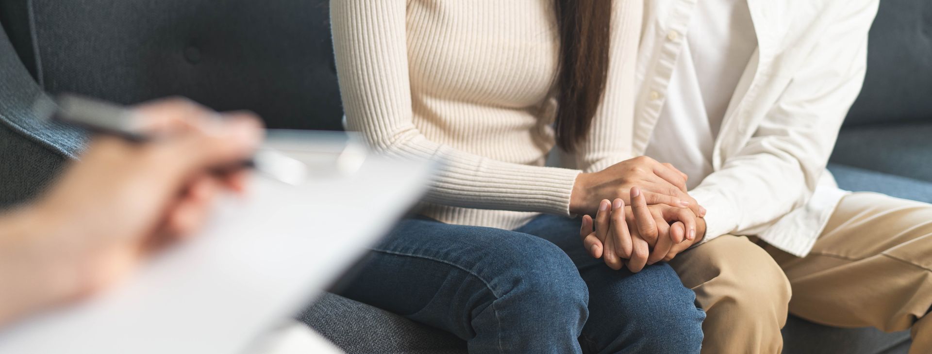 Couple holding hands during therapy session. Therapist's hand holding a pen and clipboard is in the foreground. Couple holding hands during therapy session. Therapist's hand holding a pen and clipboard is in the foreground.