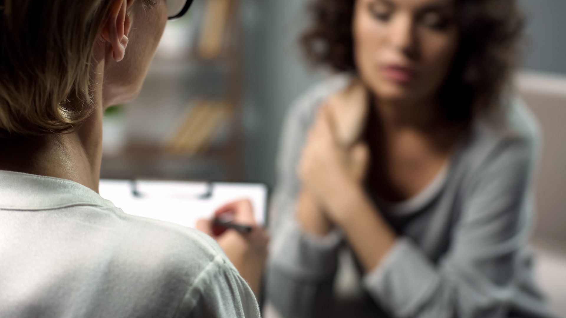 Therapist taking notes during session with a woman, who looks distressed and is holding her arms.