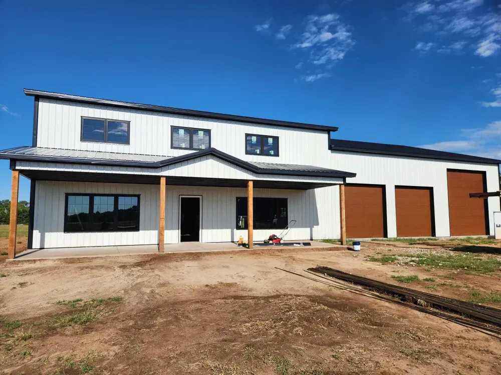 A large white house with brown garage doors is sitting on top of a dirt field.