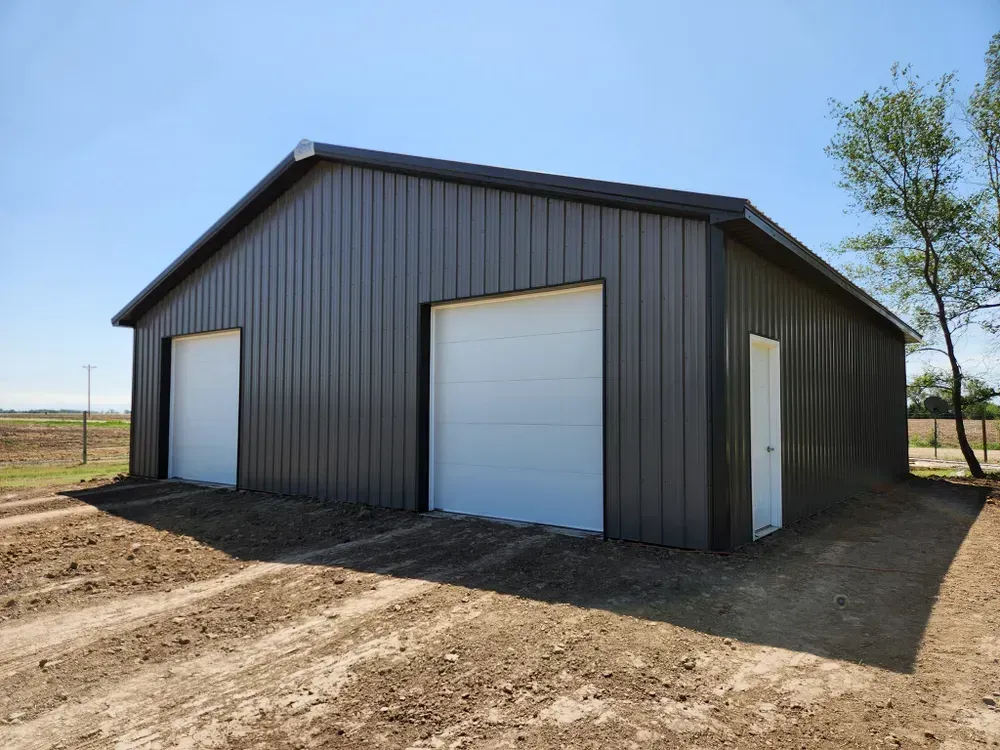 A large metal building with two garage doors is sitting in the middle of a dirt field.