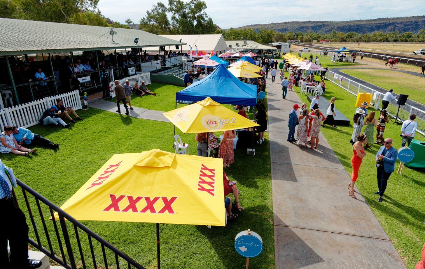 People at a sunny horse racing event with tents, lawn seating, and spectators near a track.