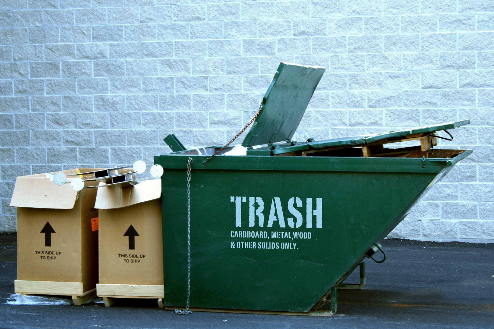 Green dumpster beside two cardboard recycling bins against a brick wall.