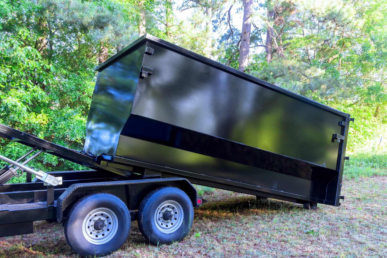 Black dump trailer with twin axles parked in a wooded outdoor area