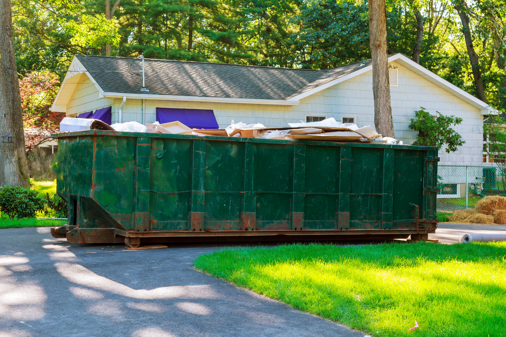 Large green dumpster filled with debris on a driveway in front of a white house and trees