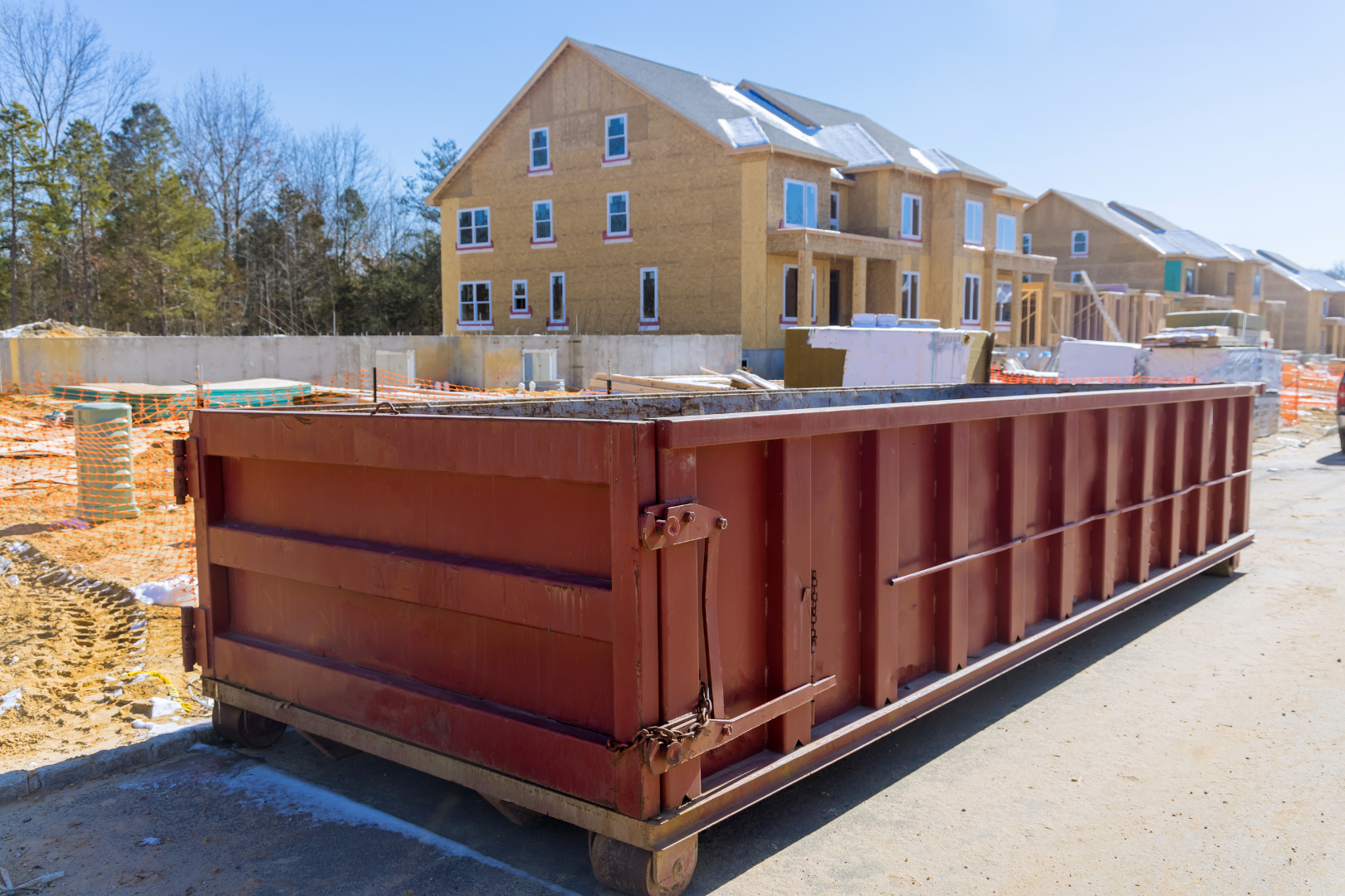Large rust-colored dumpster on a construction site beside a row of tan townhouses under construction.