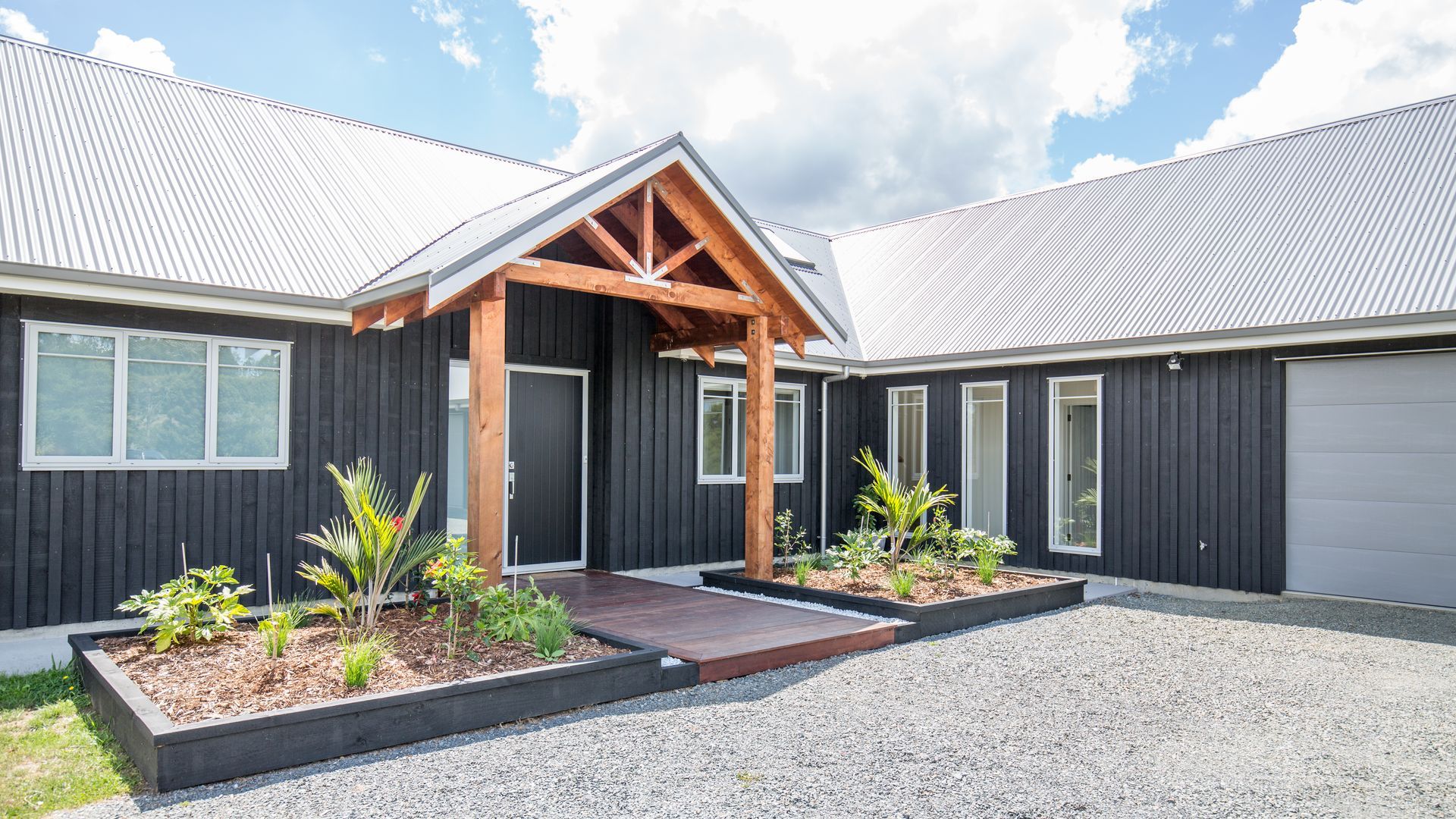 Black house with wooden entry and planters, gravel driveway, cloudy sky.