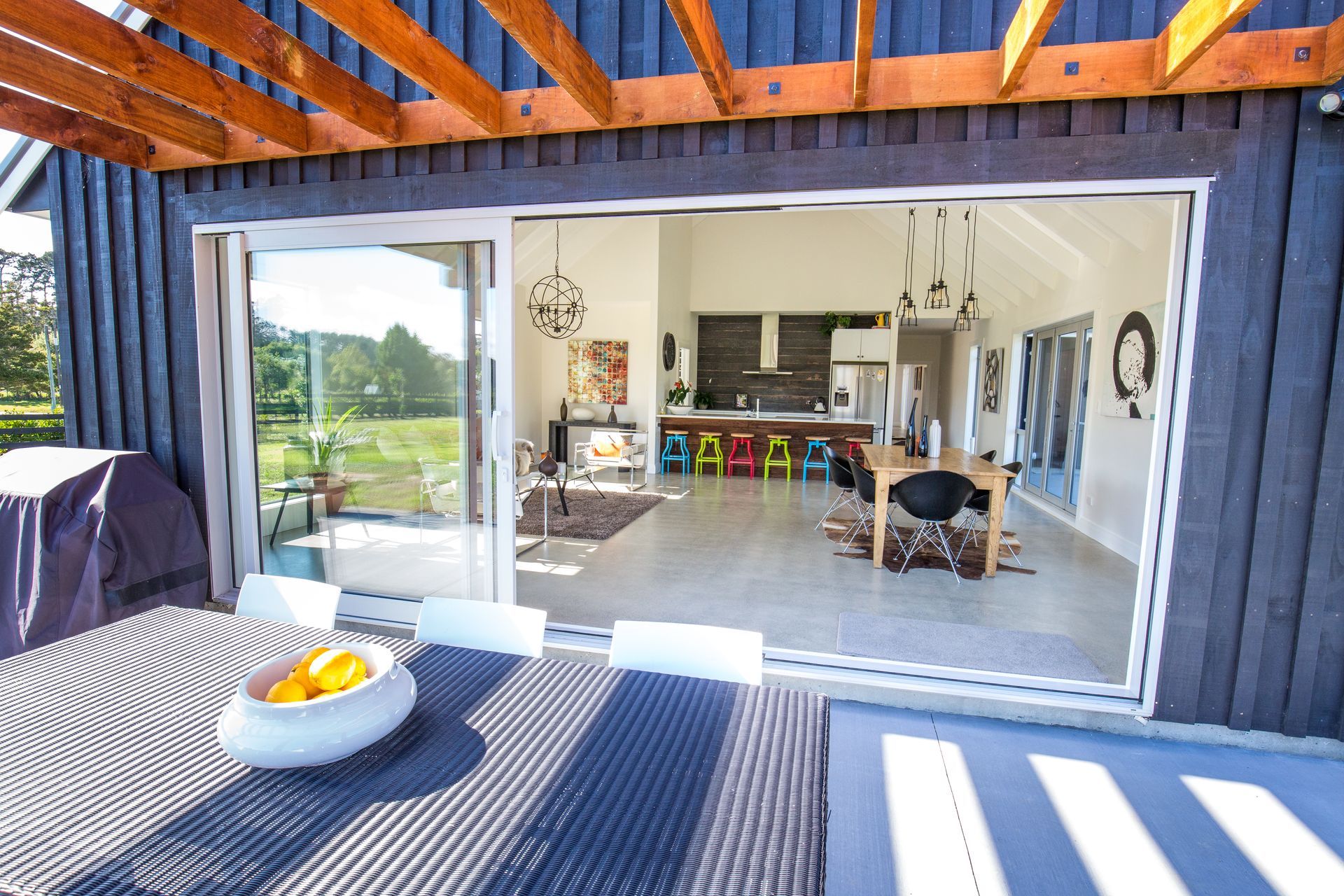 Open sliding glass doors reveal a modern home interior from an outdoor patio with a table and fruit bowl.