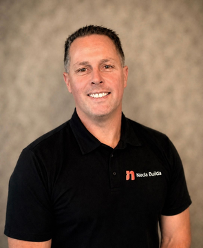 Smiling person in a black polo with a logo, standing against a neutral studio background. Andrew