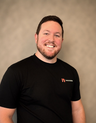 Smiling man in a black T-shirt with a small logo, standing against a beige backdrop. Brad, Project Manager.