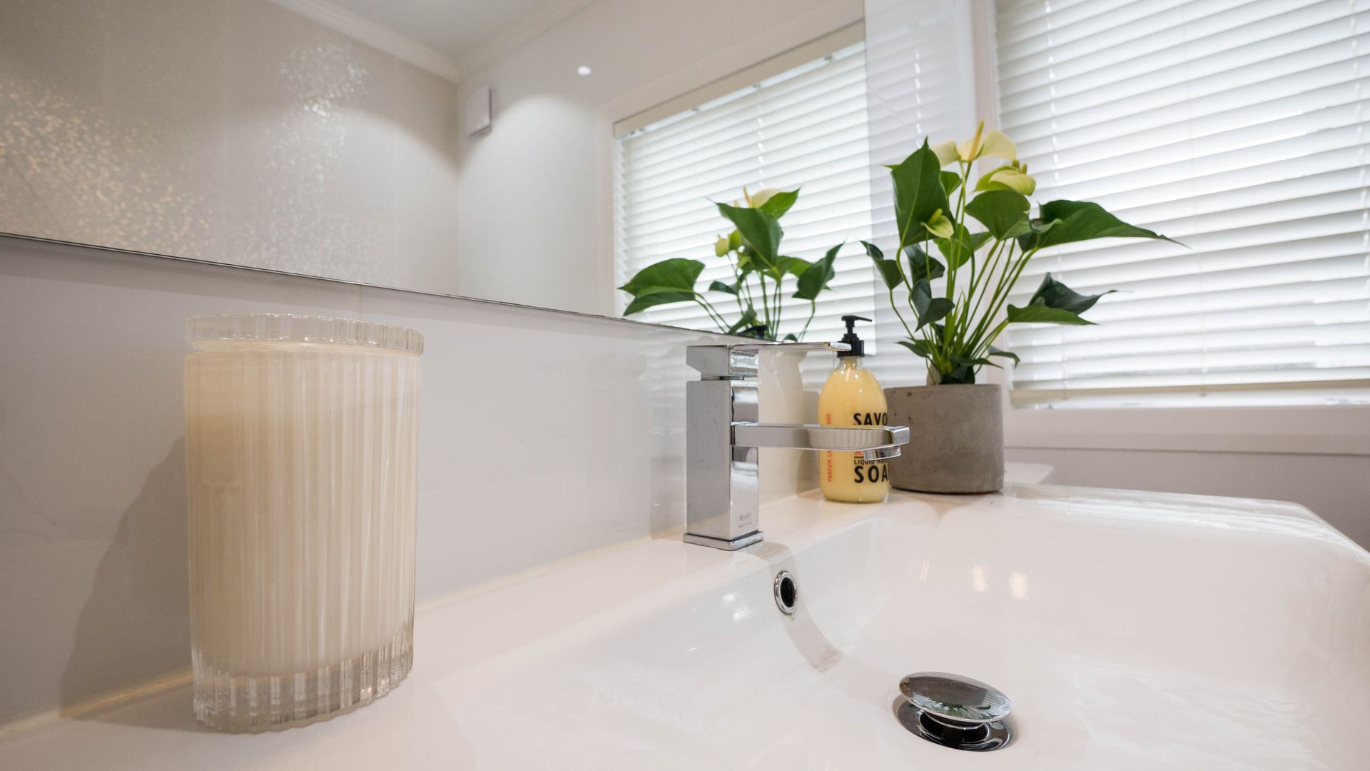 White bathroom sink with plants, soap dispenser, and candle; a mirror reflects a window with blinds.