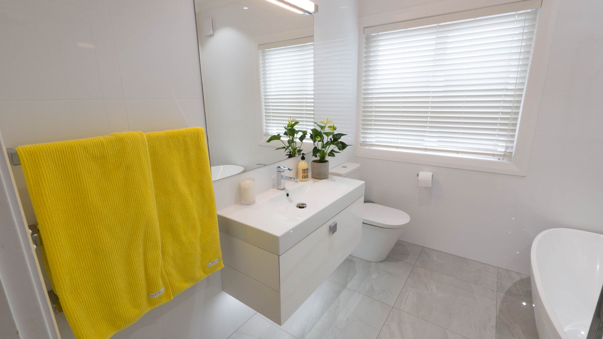 Modern white bathroom with yellow towel, floating vanity, and window with blinds.