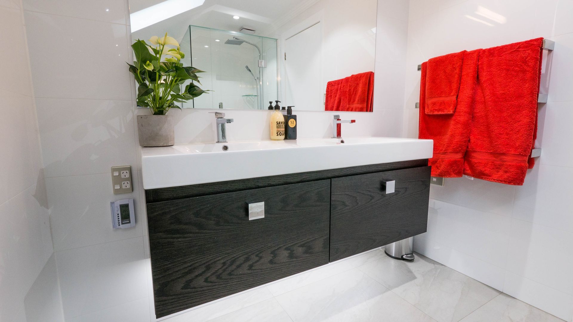Modern bathroom with a dark vanity, white counter, red towels, and a plant.