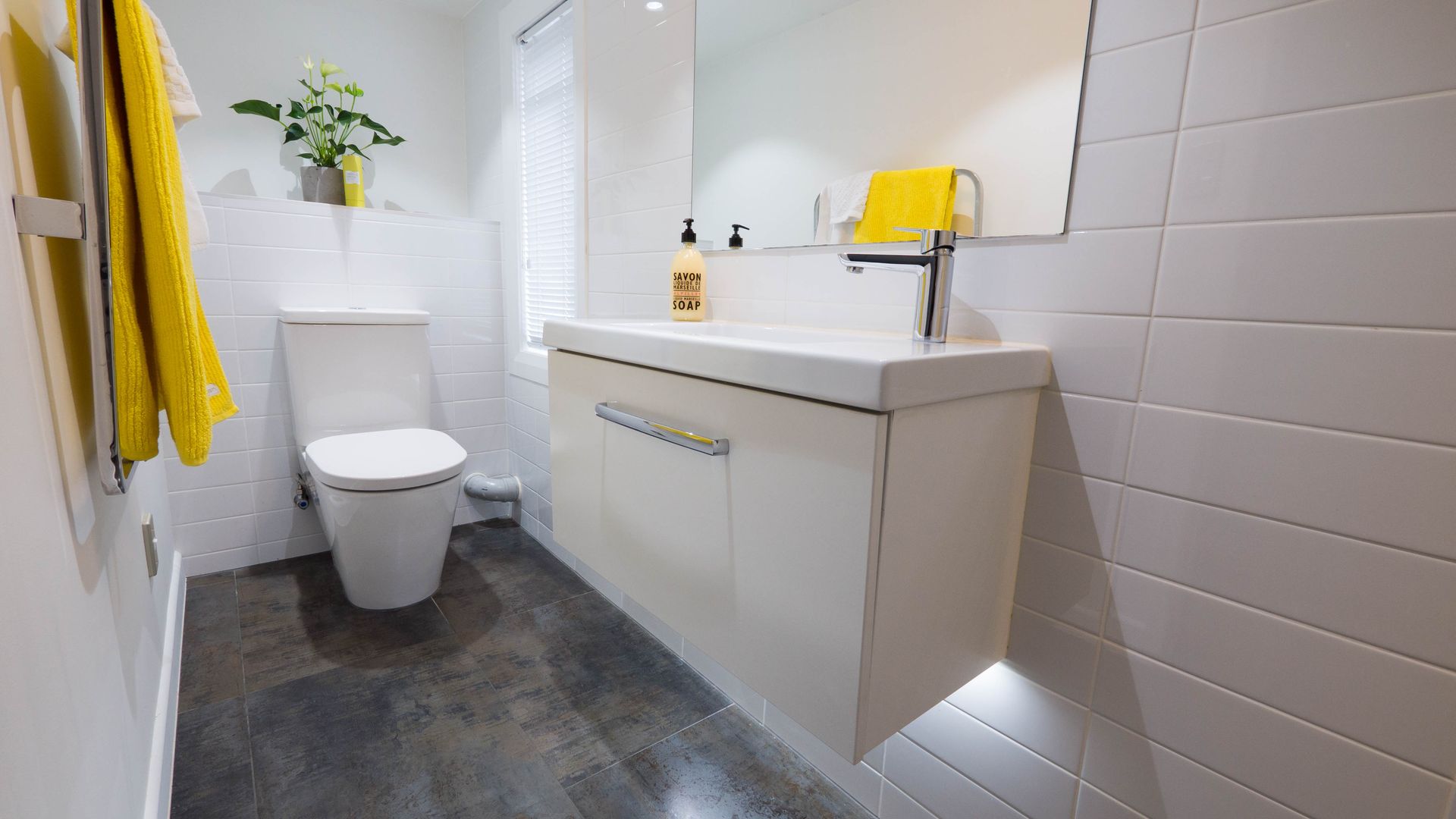 Modern bathroom with a white floating vanity, toilet, and yellow towel. Dark gray flooring and white subway tiles.