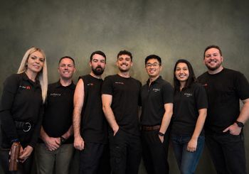 Leadership Team of 6 people standing together indoors, smiling in black shirts against a gray wall.