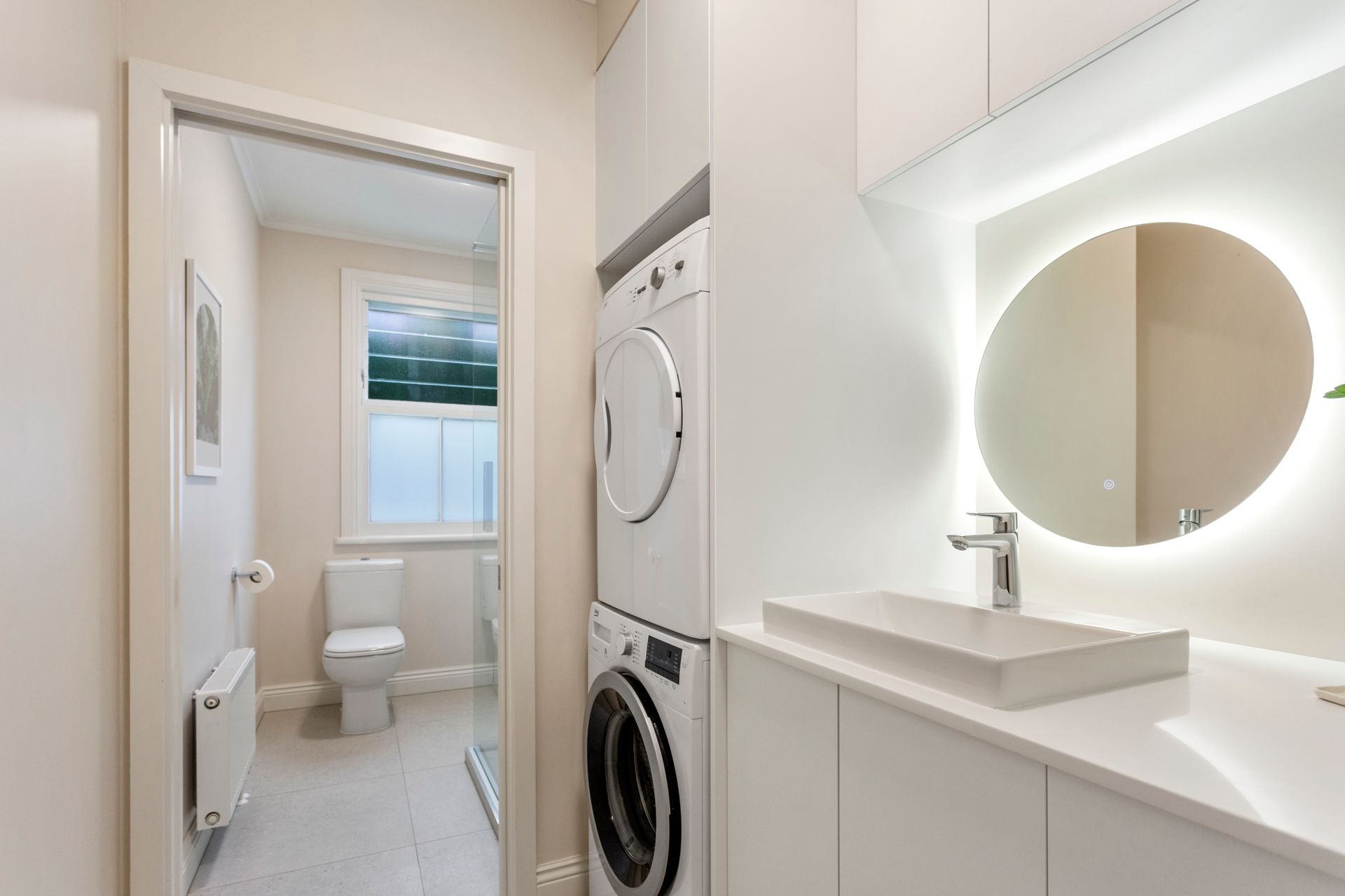 Small, modern laundry room with toilet, washer/dryer, sink, and backlit round mirror. White and neutral tones.