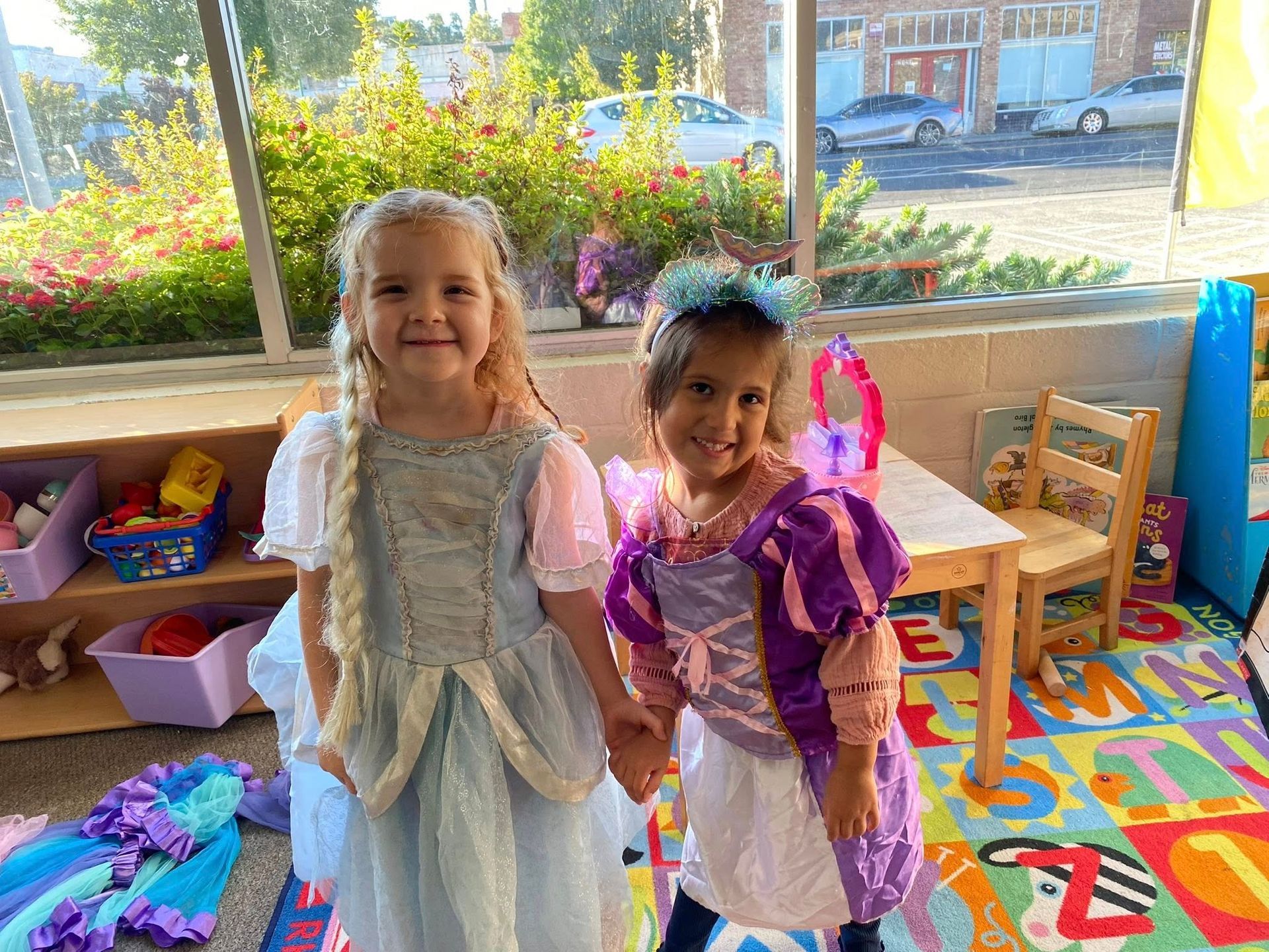 Two young children dressed as princesses, holding hands, smiling. Indoors, next to a window.