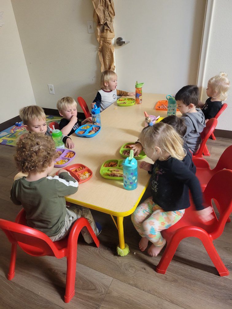 Children seated around a yellow table eating lunch; red chairs, food, and drinks visible.
