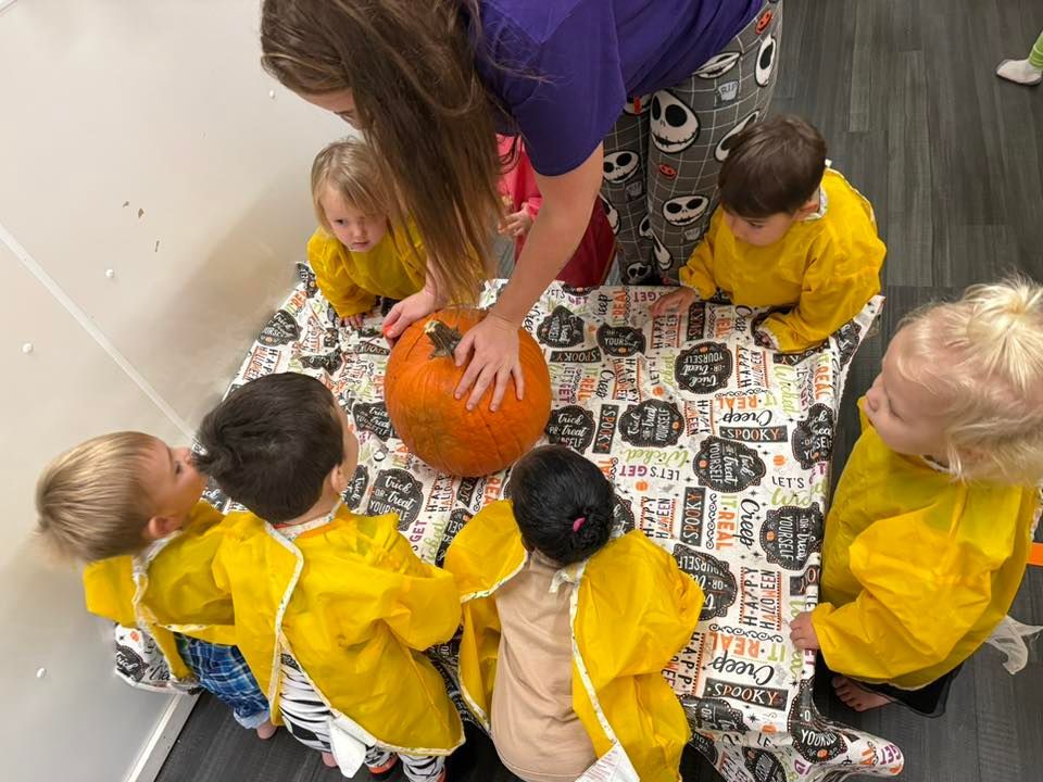 Children in yellow smocks gather around a pumpkin with an adult, all at a table.