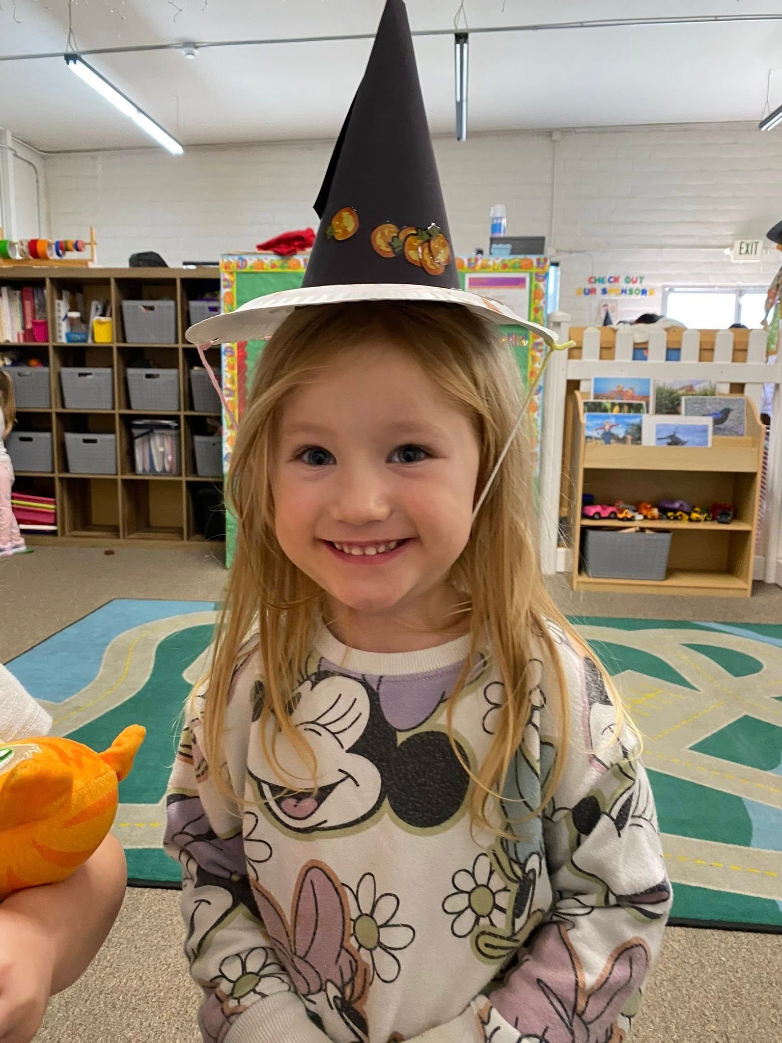 Girl wearing a witch hat smiles at the camera in a classroom, floral shirt.