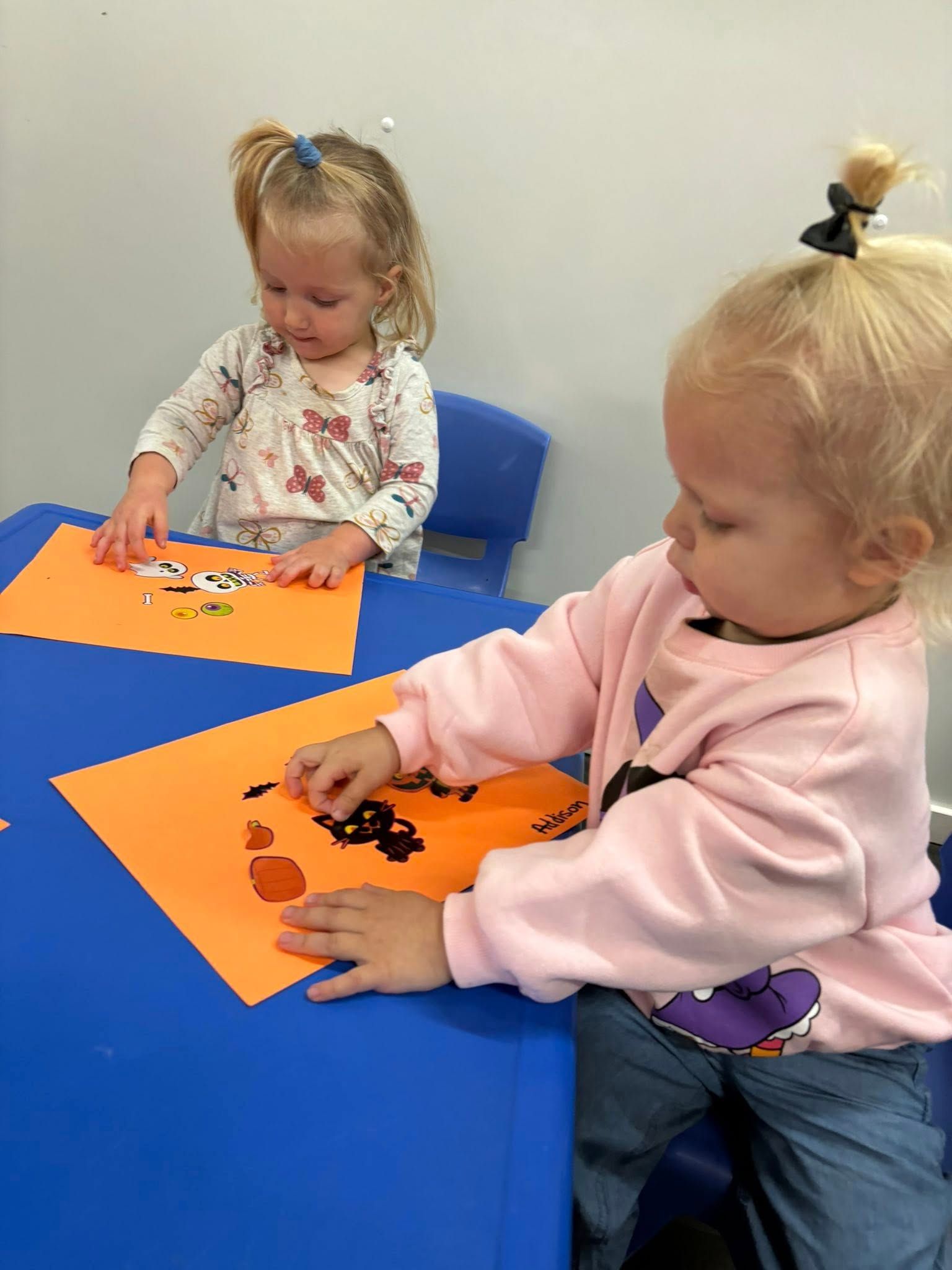 Two young children making crafts at a blue table using orange paper, black and orange items.