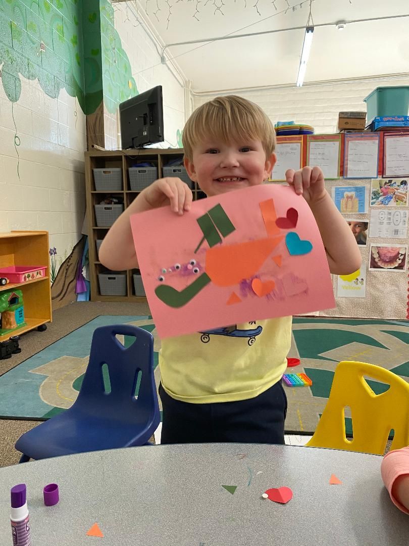 Boy in yellow shirt holding a pink paper with colorful cut-outs, smiling in a classroom.