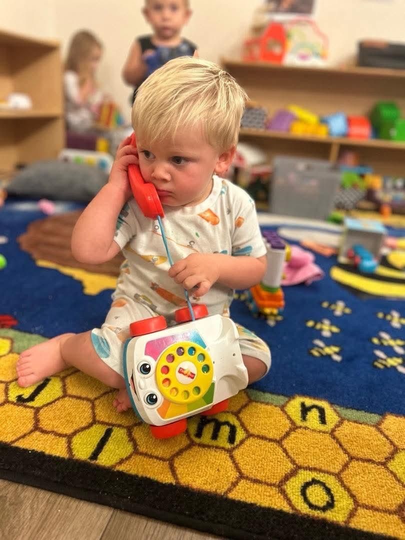 Young child sitting, holding toy telephone to ear, looking thoughtful.