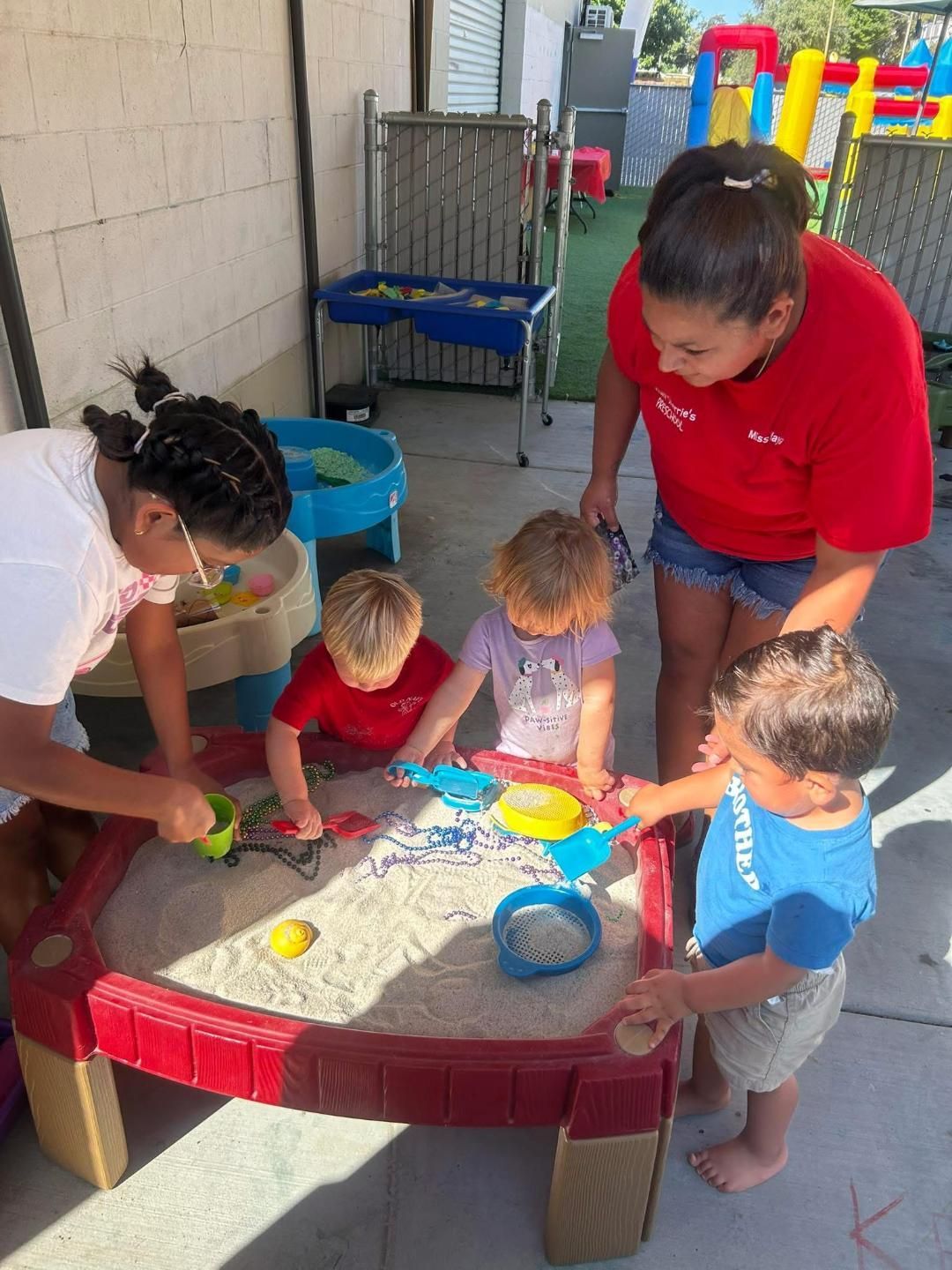 Two adults supervise children playing with sand toys at an outdoor playset.