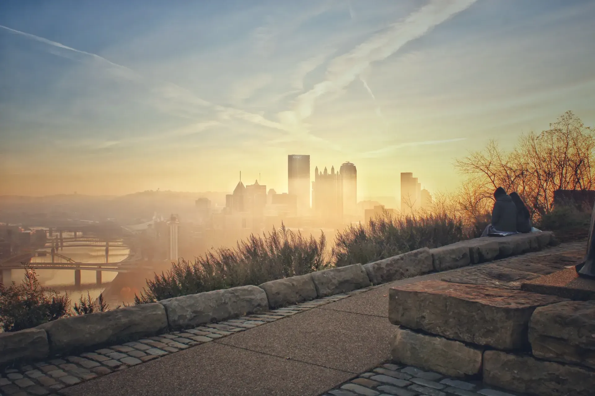 A couple is sitting on a stone wall overlooking a city at sunset.