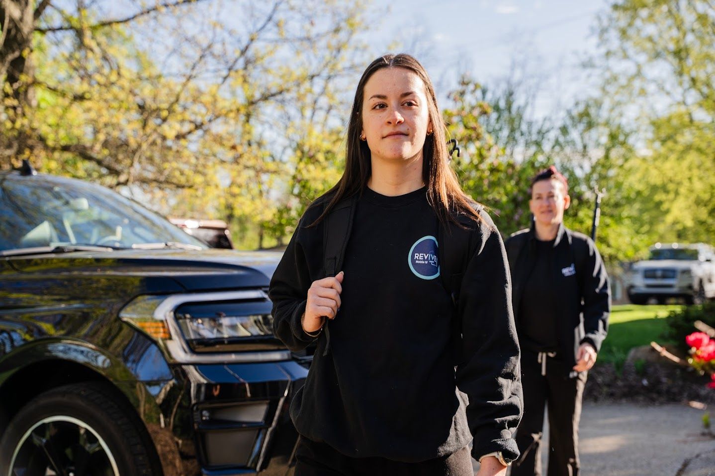A woman is standing in front of a car while a man stands behind her.
