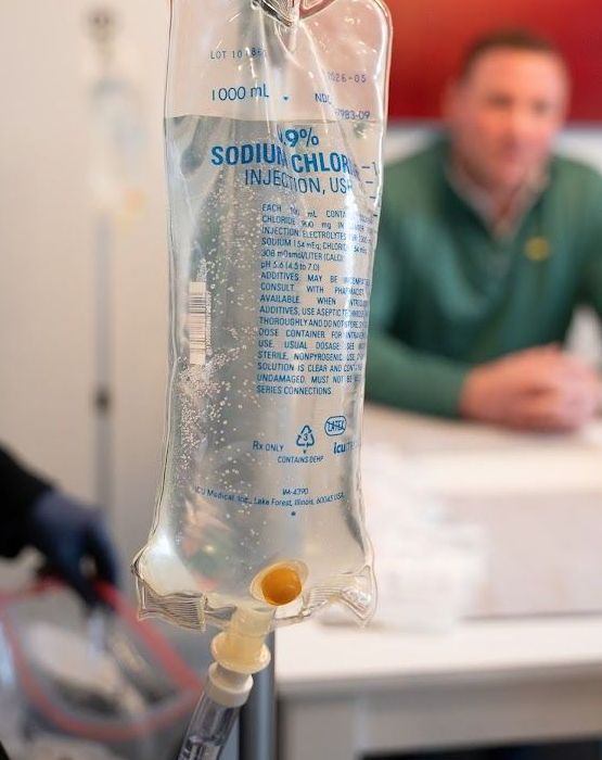A man is sitting at a table with a bag of IV liquid in front of him.
