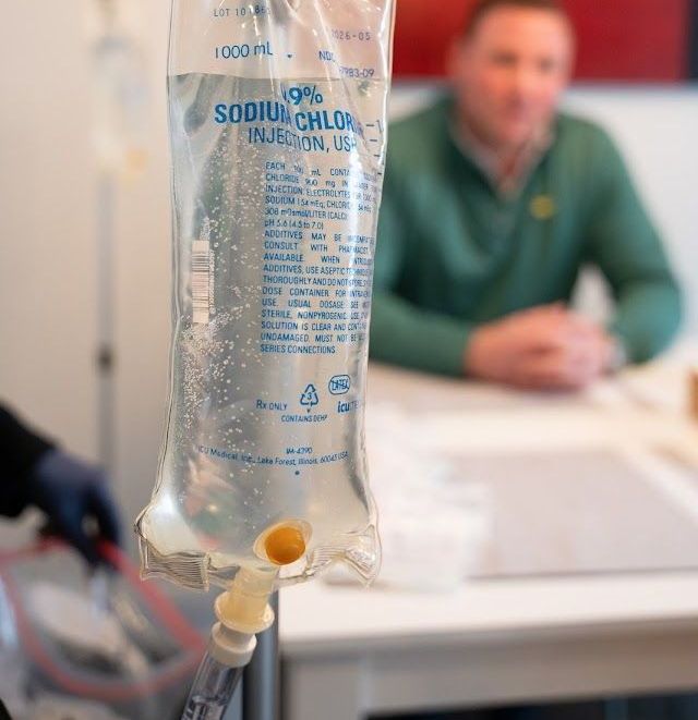 A bag of sodium chloride is hanging from a pole in front of a man sitting at a table.