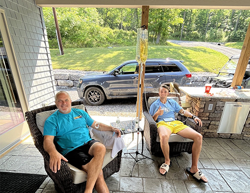 Two men are sitting on a porch with a car parked in the background.