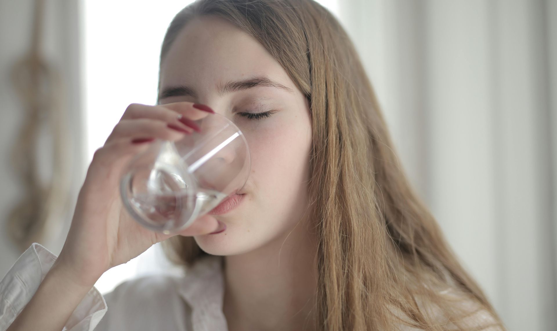 Woman with long blonde hair drinking water from a glass, eyes closed.