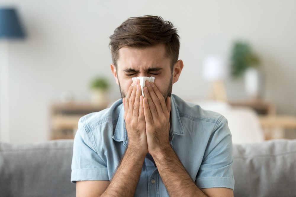 A man is blowing his nose into a napkin while sitting on a couch.