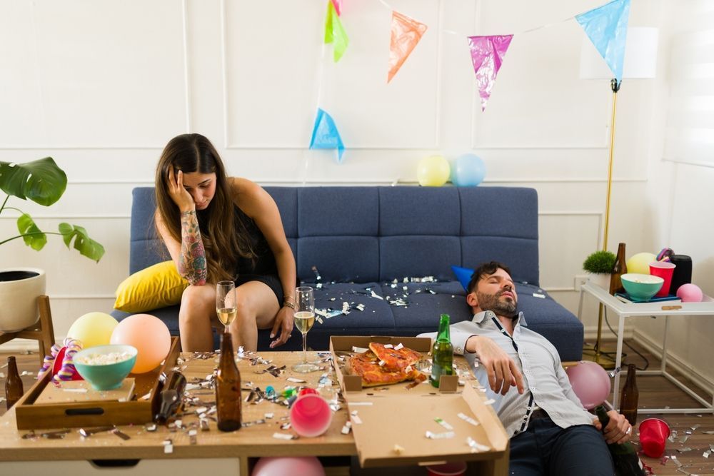 A man and a woman are sitting on a couch in a messy living room after a party.