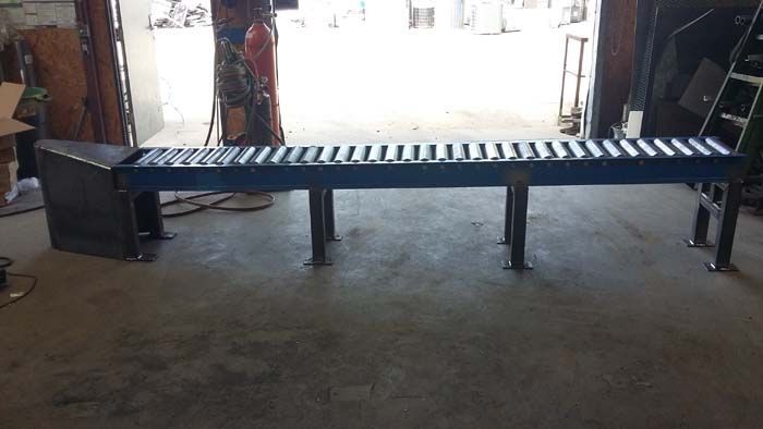 A long, blue industrial roller conveyor table sits on metal legs in a workshop, ending with a triangular steel side panel.