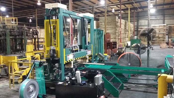 A large green and yellow industrial pallet strapping machine sits on a concrete floor in a warehouse with lumber stacks.