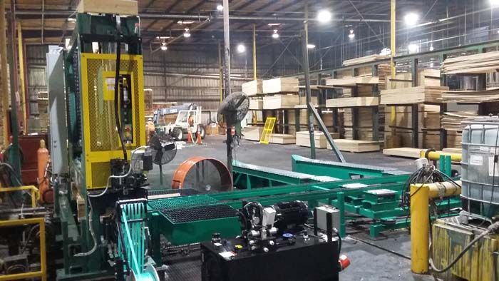Industrial machinery with a yellow safety cage stands in a large warehouse filled with stacked lumber on storage racks.