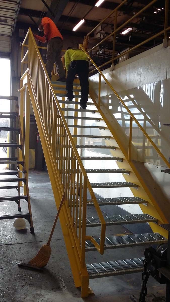 Two workers in high-visibility vests climb a yellow metal industrial staircase inside a warehouse.