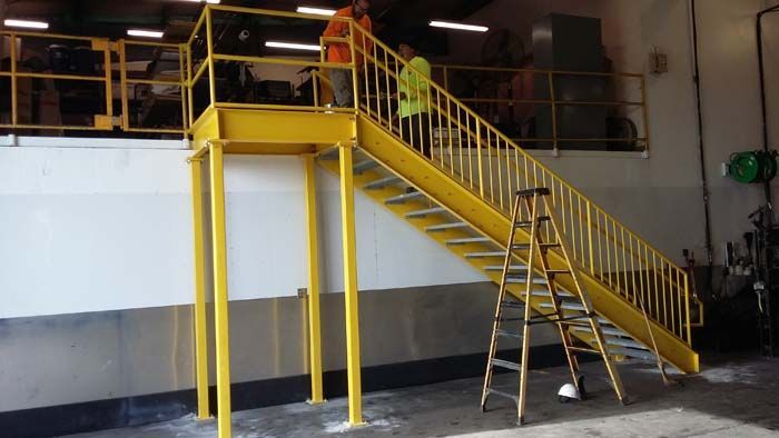 Yellow metal stairs and an elevated platform in an industrial space, with two individuals standing on the landing.