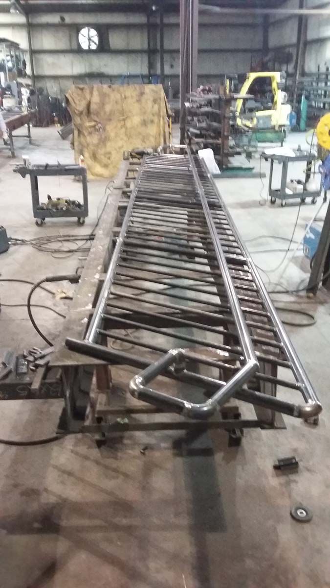 A long metal railing with a decorative, angular handle lies on a workbench inside a metal fabrication workshop.